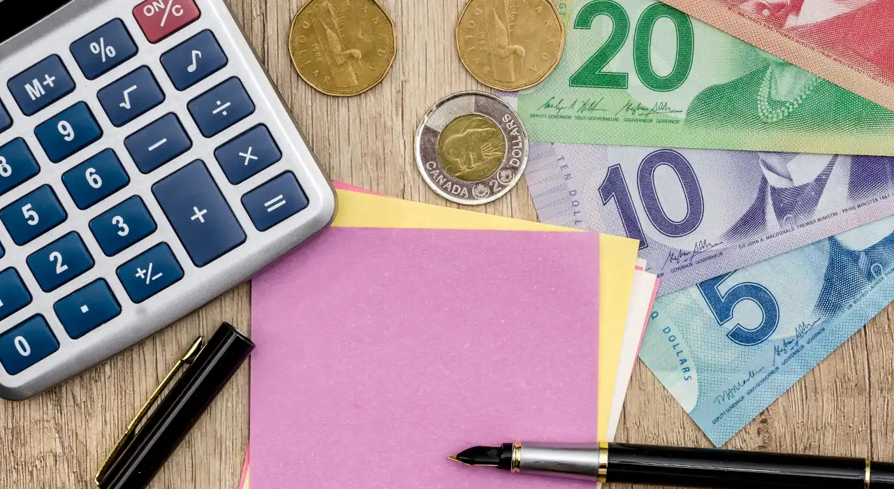 ਮੇਜ਼ ‘ਤੇ ਕੈਲਕੂਲੇਟਰ, ਕੈਨੇਡੀਅਨ ਸਿੱਕੇ ਅਤੇ ਨੋਟ, ਨਾਲ ਹੀ ਕਲਮ ਅਤੇ ਸਟਿੱਕੀ ਨੋਟ।