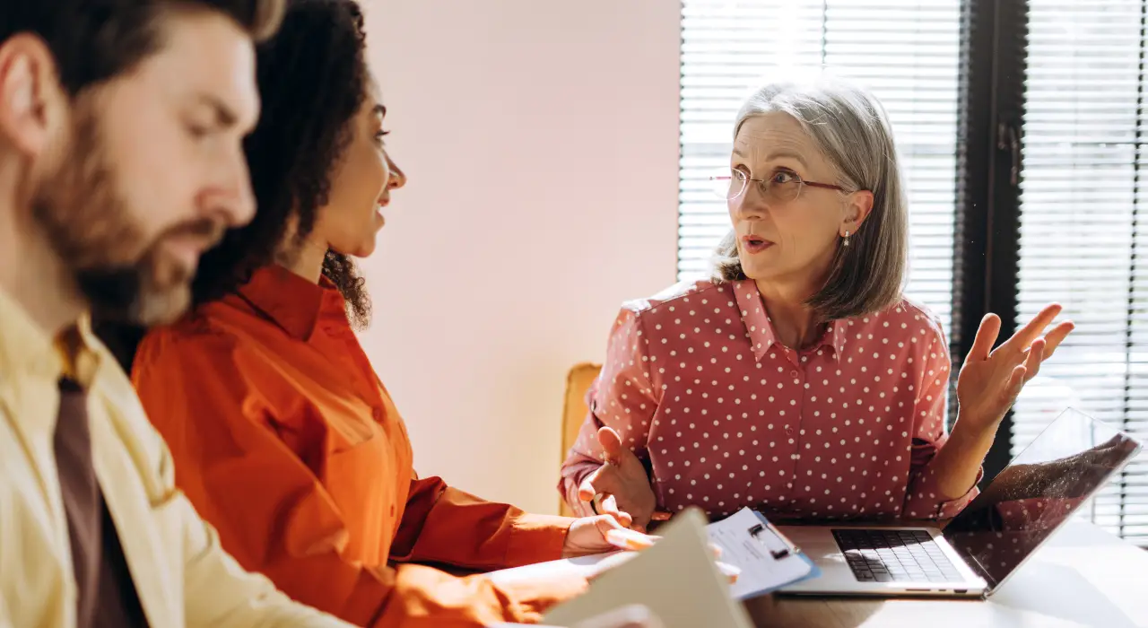 Trois personnes assises à une table avec des ordinateurs portables et des documents, engagées dans une discussion sérieuse.