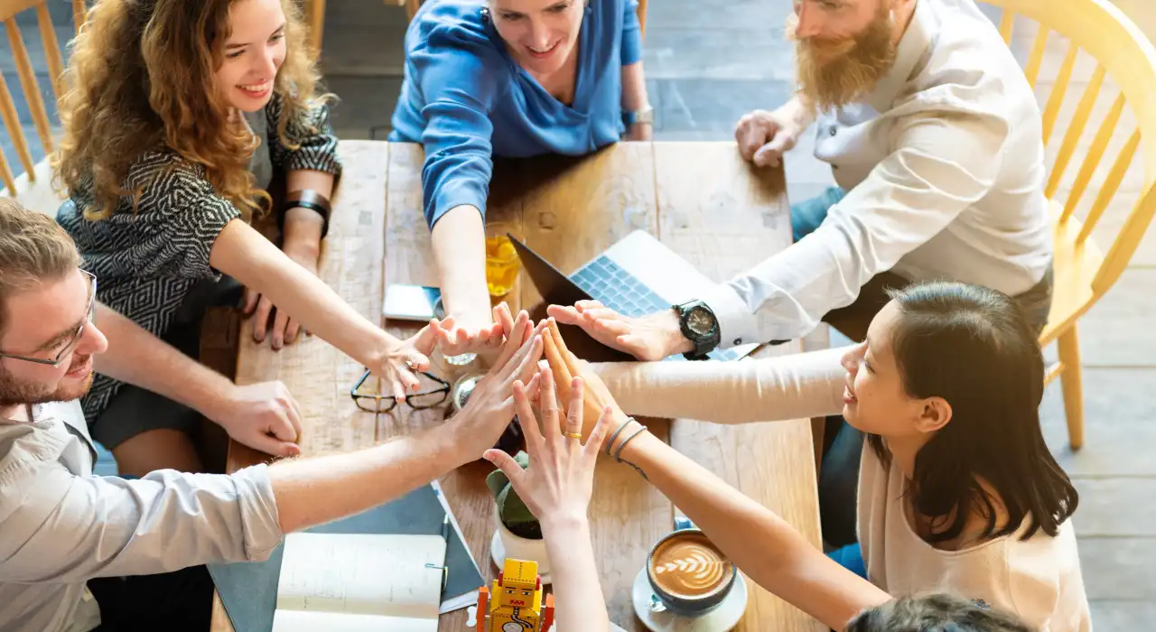 ਛੇ ਲੋਕ ਲੱਕੜ ਦੀ ਮੇਜ਼ ਦੇ ਆਲੇ-ਦੁਆਲੇ ਬੈਠੇ, ਮੁਸਕਰਾਂਦੇ ਹੋਏ, ਟੀਮ ਦੇ ਇਸ਼ਾਰੇ ਵਜੋਂ ਆਪਣੇ ਹੱਥ ਇਕੱਠੇ ਰੱਖਦੇ ਹੋਏ।