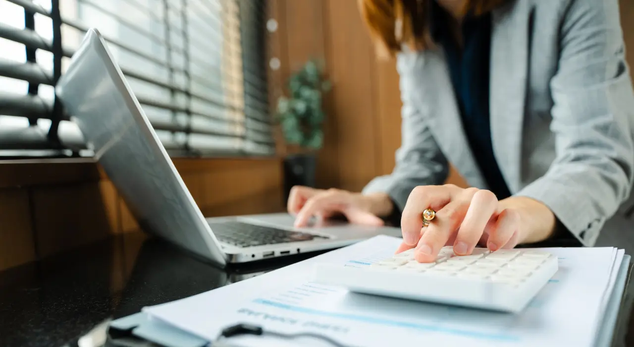 Una persona usa una calculadora y una computadora portátil en un escritorio con documentos financieros a la vista.