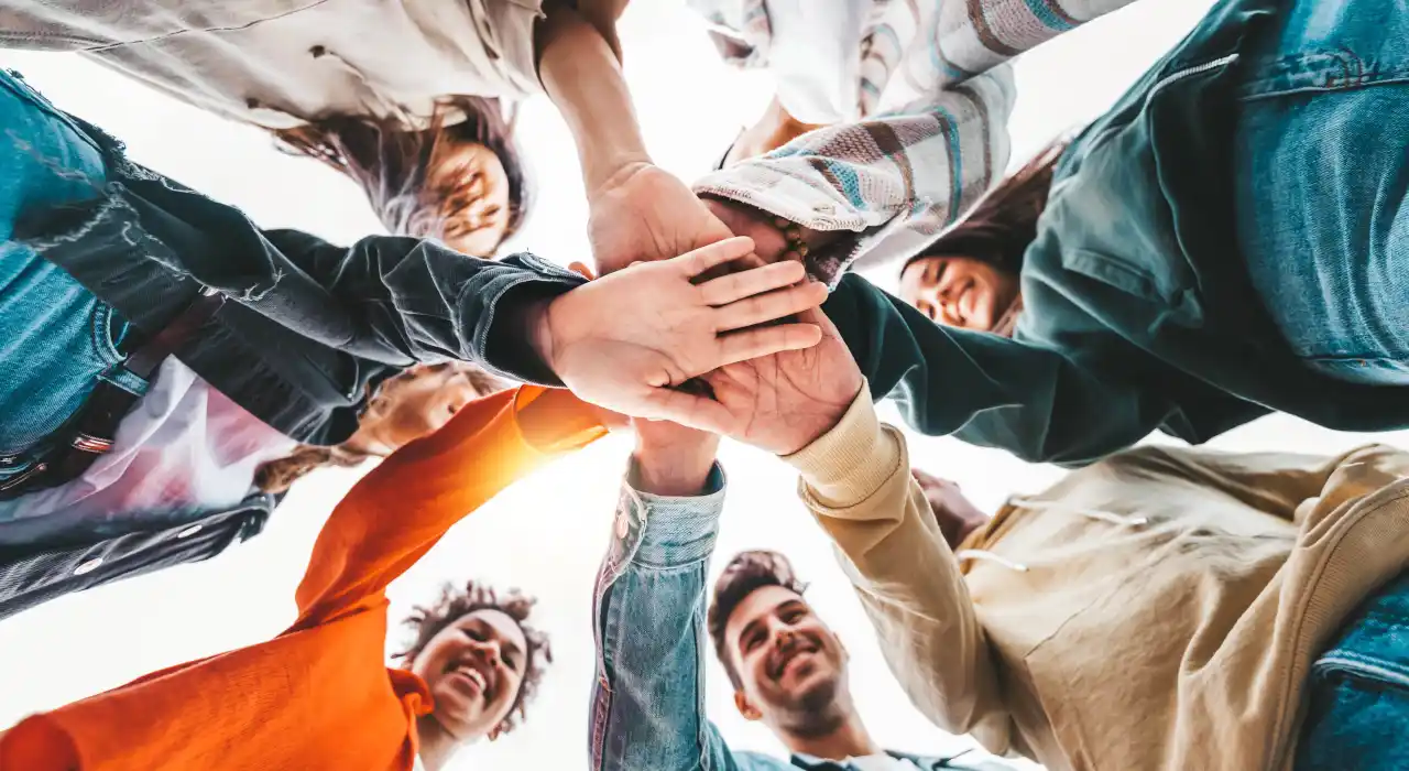 ਵੱਖ-ਵੱਖ ਪਿਛੋਕੜਾਂ ਦੇ ਨੌਜਵਾਨਾਂ ਦਾ ਸਮੂਹ ਗੋਲ ਦਾਅਰਾ ਬਣਾਕੇ ਖੜ੍ਹਾ ਹੈ, ਮੁਸਕਰਾਂਦੇ ਹੋਏ, ਹੱਥ ਇਕੱਠੇ ਕਰਦੇ ਹੋਏ।